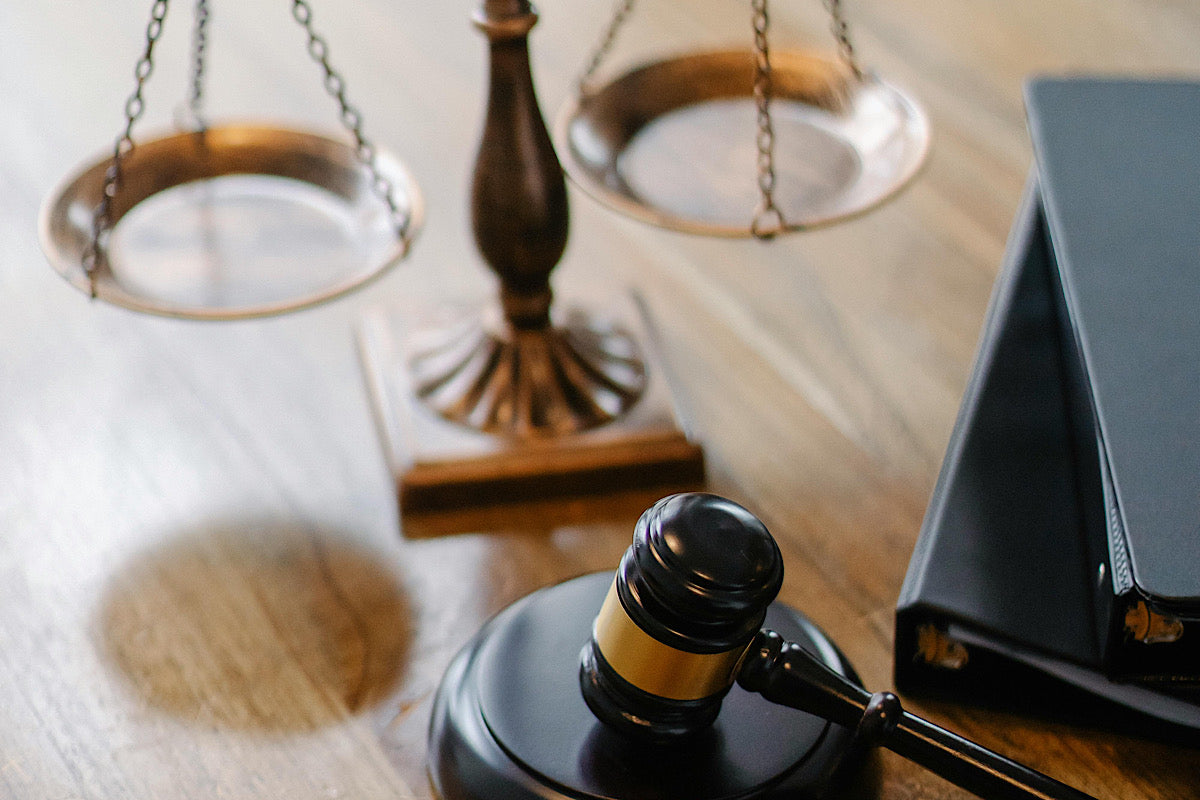 An ebony, wooden gavel with a gold band around the head is shown with one strike surface resting on the gavel base and in front of a set of copper scales of justice