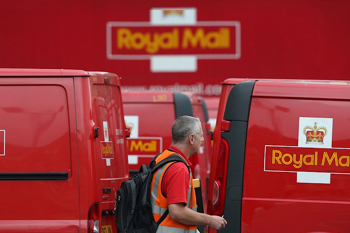 A man in a red Royal Mail polo shirt, orange hi-vis jacket and wearing a black rucksack is captured central to the image as he walks from left to right. Behind him, a fleet of Royal Mail vans are parked in a Royal Mail depot, lined up in two rows back to back. The backdrop is a large Royal Mail logo on a Royal Mail Red surface.